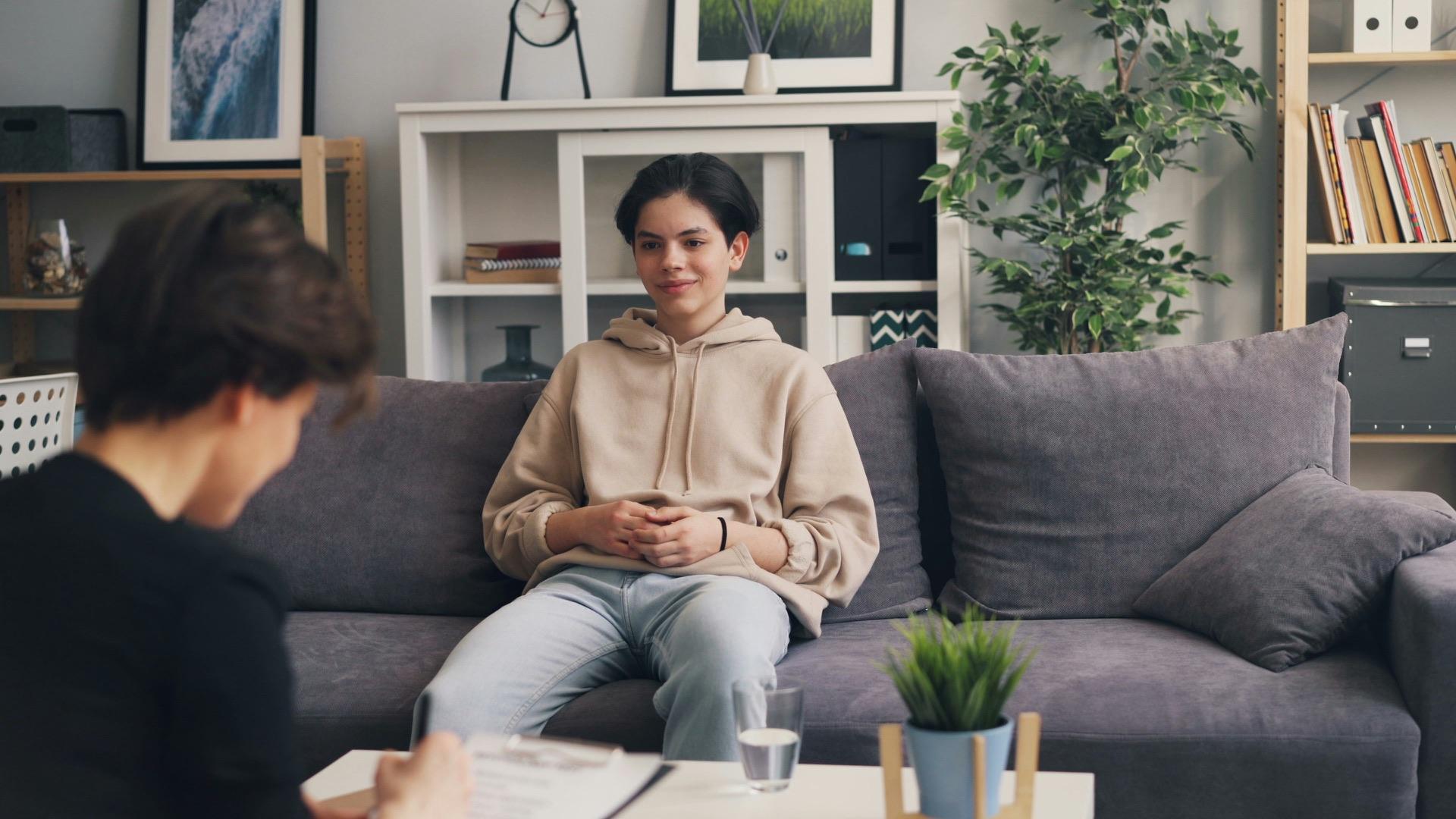 A young person in a beige hoodie in a lived-in front room, in quiet conversation with a key worker across a low table.
