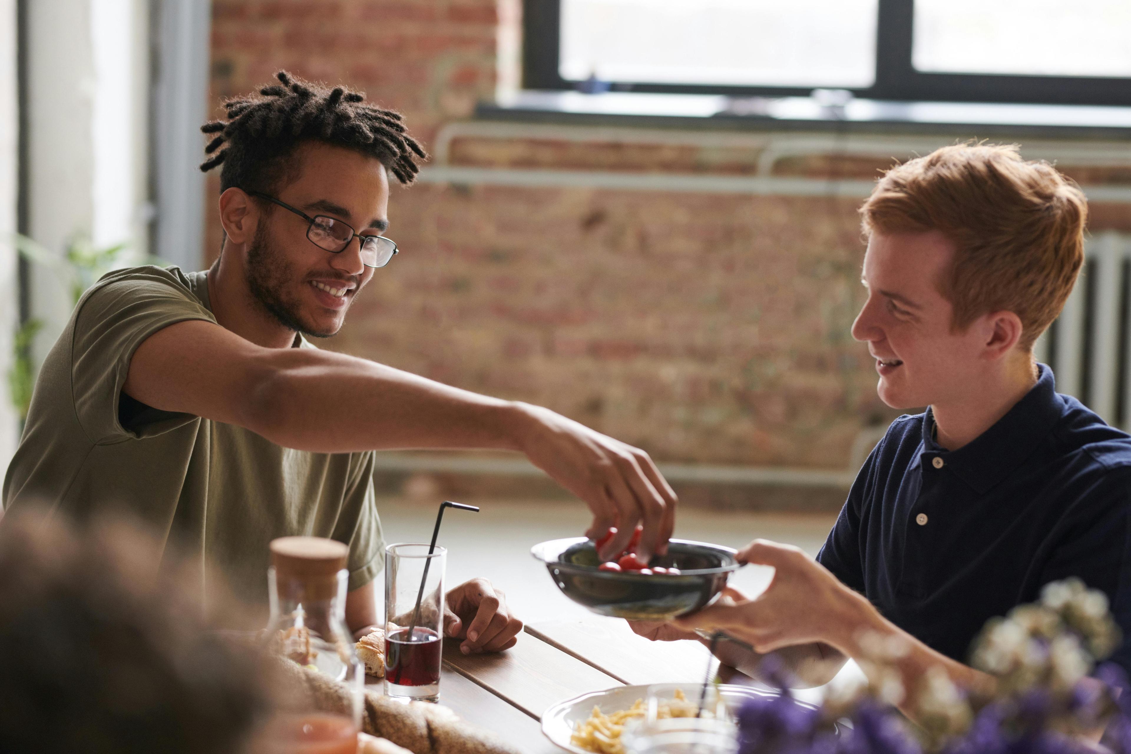 Two young men sharing a casual meal at a sunlit table, one passing a bowl to the other.